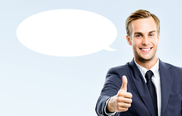 Portrait image - happy smile business man wear dark suit jacket, tie showing pointing thumb up hand sign gesture. Businessman isolated grey gray background with white empty mockup speech bubble