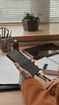 Vertical closeup of unrecognizable office employee connecting powerbank to her smartphone using cable while sitting at wooden desk covered with paperwork