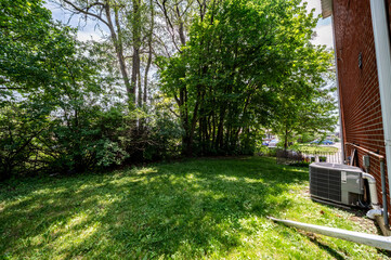 Obraz premium Lush green lawn and mature trees fill a residential backyard on a bright day, featuring a central air conditioning unit positioned against a brick wall of a suburban home