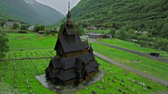 Aerial close view of Borgund Stave Church roof and cemetery, Norway. Dark wooden structure contrasts with green grass and surrounding hills.