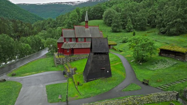 Aerial view approaching Borgund Stave Church in Norway. Medieval wooden structures and stone paths are surrounded by lush hills and forest.