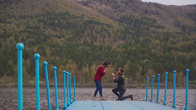 Young man proposing to girlfriend with wedding ring on the pier with beautiful view on mountain lake. She said yes. Surprise engagement.