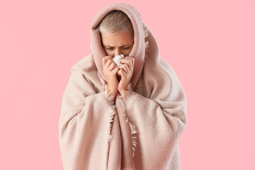 Sick mature woman with blanket and tissue sneezing on pink background