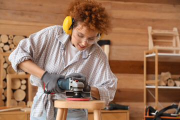 Young female carpenter polishing wooden stool with angle grinder in workshop