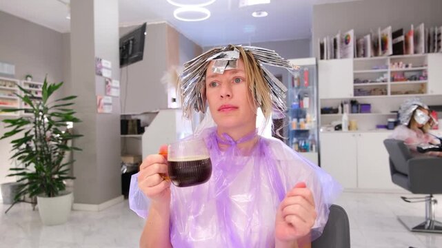 Moscow, Russia, 24.04.2025 Woman with hair in foil highlights drinking coffee, relaxing during hair treatment in beauty salon footage