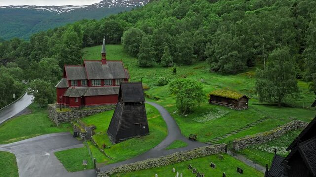 Drone view of a historic church, bell tower, and nearby red buildings. Borgund Stave Church lies among rolling hills and rural roads.