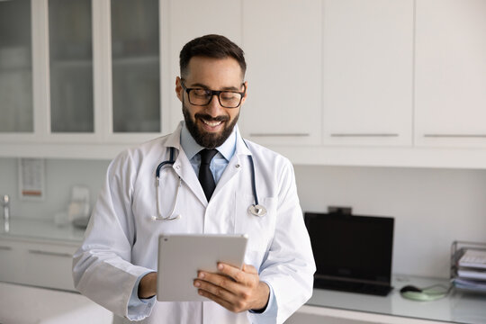 Digital healthcare professional. Smiling male doctor in white hospital coat use electronic pad in clinic office review medical records therapy plans lab results contact with patient via telehealth app