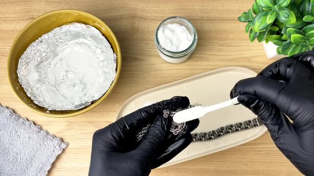 Cleaning silver jewelry with a brush and baking soda. Gloves are used, and the photo is taken on a wooden table at home.