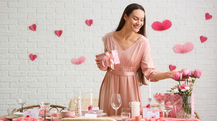 Young woman with gift and roses near festive table setting in dining room. Valentine's Day...
