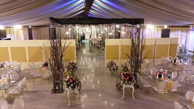 A smooth aerial drone shot capturing the grand entrance of a luxury wedding marquee featuring a floral-decorated walkway, elegant hanging lights, and symmetrical event styling. 