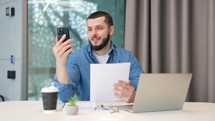 Professional adult engages in happy video call, reviewing documents. Modern office workspace features laptop and coffee, highlighting successful virtual communication and remote work efficiency.