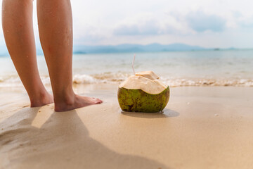 Barefoot person standing on sandy beach near fresh green coconut with straw, calm sea and distant mountains creating tropical vacation scene 