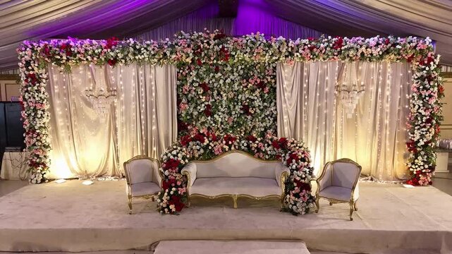 A cinematic aerial drone shot capturing an elegant wedding stage decorated with beautiful floral arrangements and soft ambient lighting. The scene highlights a romantic and luxurious atmosphere, 