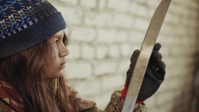 Dirty kid in wool cap and gloves holding cutlass thoughtfully near brick wall, face smeared with dust, gazing at blade with serious expression in worn jacket showing hardship and resilience
