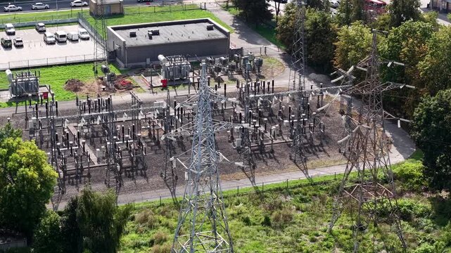 Drone aerial over a high-voltage electrical substation with switchgear, transformers and transmission towers beside urban greenery on a clear day.