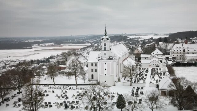 4K drone pullback reveals snowy church tower, cemetery and village. Smooth widening captures elegant winter composition&mdash;ideal for promos, documentaries, tourism visuals.