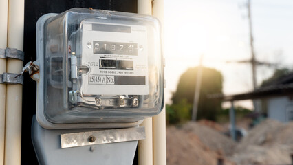 Analog electric meter encased in a transparent glass or plastic housing. Digital-style rolling counters display the energy consumption digits. Mounted on a vertical support with conduit pipes. © thongchainak