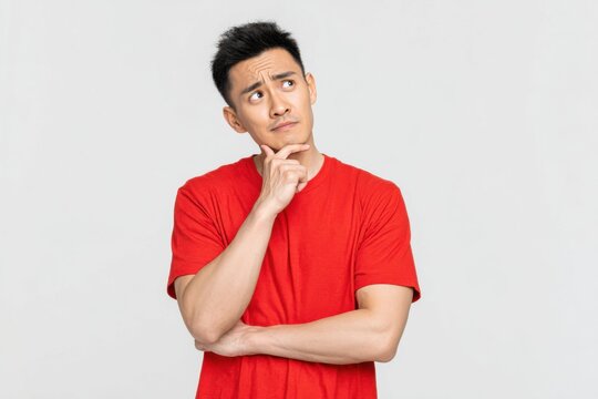 Young Asian man wearing a red shirt stands in a neutral setting, contemplating and expressing curiosity about a thought or idea during a bright afternoon