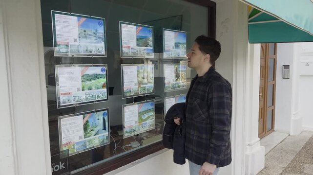 A man stands outside a real estate agency in Spain looking at property listings displayed in the window, showcasing local housing opportunities.