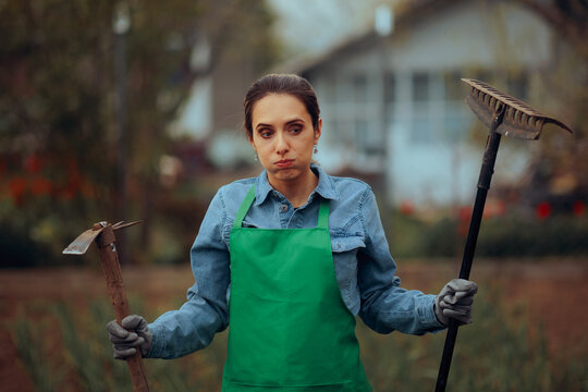 Puzzled Farm Worker Holding a Hoe and a Rake. Clueless female farmer standing with tilling equipment in hand 
