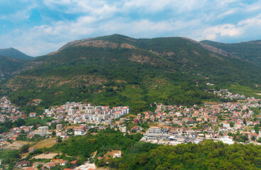 Obraz premium Aerial view of a coastal town in Montenegro nestled at the foot of lush green mountains under a blue cloudy sky.