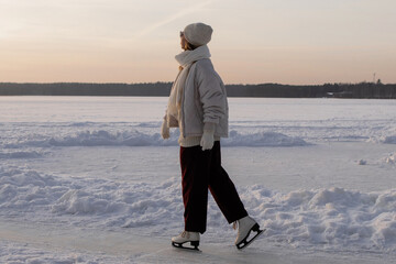 Silhouette of woman ice skating on frozen lake at golden sunset with dramatic orange sky and forest...