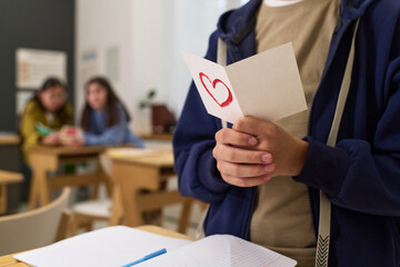 Teenage boy with light skin holding handmade card with red heart drawing in classroom, standing near desk with open notebook, two teenage girls sitting and talking in background