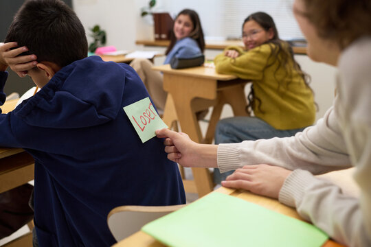 Group of multiethnic teenagers sitting in classroom, Caucasian teenage girl secretly placing sticky note with loser written on back of boy while others watching and smiling
