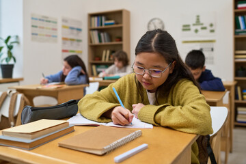 Asian preteen girl sitting at desk writing in notebook in classroom with other children studying in background, focused expression, school supplies on table, educational setting © pressmaster