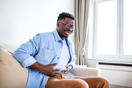 African American Man Suffering from Stomach Ache and Abdominal Pain on Sofa, Black Male Holding Belly Feeling Gastritis Symptoms or Food Poisoning, Digestive Health Problem and Cramps at Home
