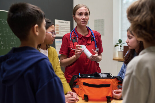 Caucasian middle aged woman paramedic demonstrating medical equipment to group of diverse children in classroom setting, children attentively listening and observing emergency kit demonstration
