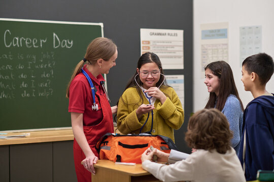 Caucasian female young adult doctor demonstrating medical equipment to diverse group of teenagers in classroom, teenagers interacting with stethoscope and smiling during educational activity