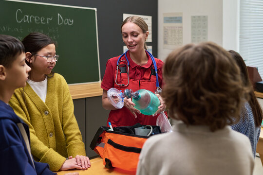 Caucasian young adult woman paramedic demonstrating medical resuscitator mask to diverse group of teenagers in classroom during career day, students attentively observing demonstration