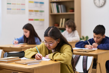 Group of multiethnic preteen children sitting at desks writing in notebooks during classroom activity, focused expressions, school supplies and books visible on desks, educational setting
