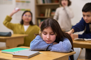 Caucasian preteen girl sitting at desk with head resting on arms appearing sad, while classmates in background interacting and one boy reaching toward her in classroom setting