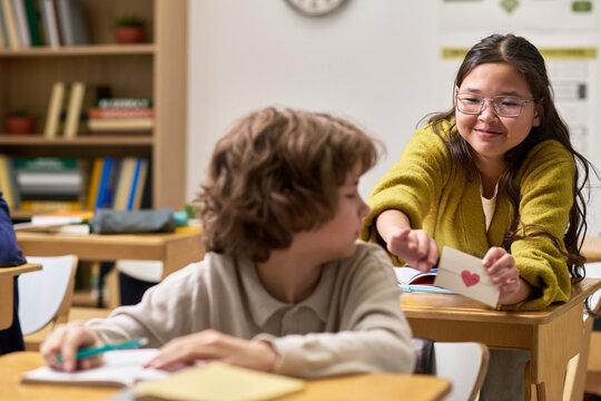 Teenage Caucasian girl smiling while handing note with heart drawing to teenage Caucasian boy sitting at desk in classroom, boy looking at note, both engaged in school activity