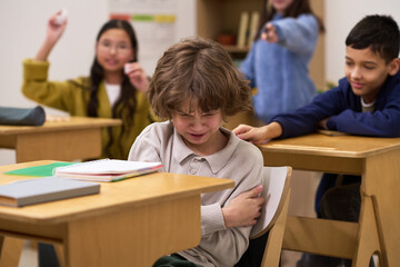 Caucasian child sitting at desk crying while multiethnic group of children in classroom pointing and laughing, showing bullying behavior during school lesson