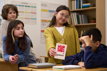 Group of multiethnic children interacting in classroom, Asian girl presenting handmade card with red hearts to boy sitting at desk, two other girls observing and smiling nearby