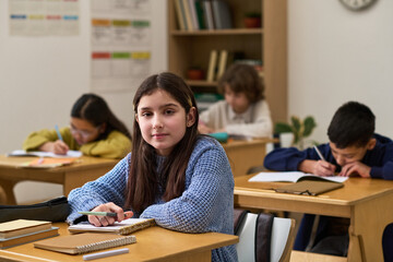 Portrait of Caucasian preteen girl sitting at desk in classroom looking at camera while classmates of diverse ethnicities studying in background writing in notebooks
