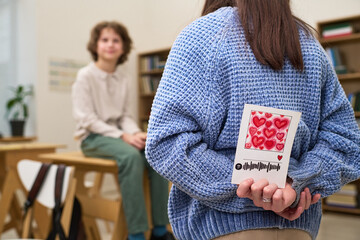 Teenage Caucasian girl hiding greeting card decorated with red hearts behind back while standing in classroom, seated teenage Caucasian boy looking at her and smiling in background