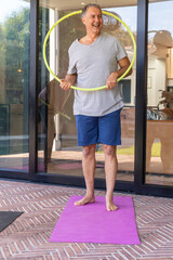 Mature man standing on purple exercise mat on brick patio, holding neon hula hoop © wavebreak3