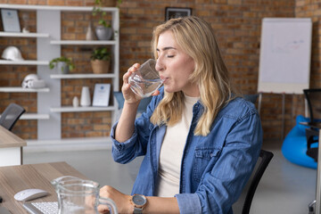 Female office worker sitting at desk drinking water from clear glass near keys wearing denim shirt