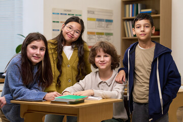 Group of multiethnic children smiling and posing together in classroom, two girls and two boys standing and sitting around desk with books, looking at camera, showing friendship