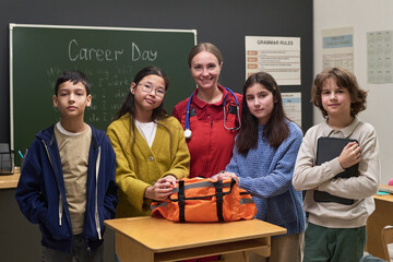 Group of diverse children standing with young Caucasian woman doctor in classroom, children posing together near desk with medical bag, blackboard in background displaying Career Day