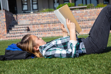 Teenage girl lying on grass and using backpack as pillow and reading paperback book in garden