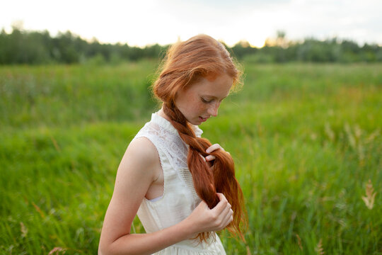 Teenager with red hair and freckles braiding hair outdoors in summer park