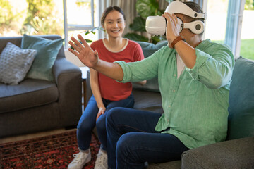 Asian couple sitting on sofa in living room using white vr headset reaching and watching