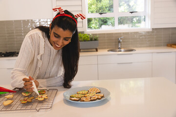 Indian woman in her thirties decorating sugar cookies on white kitchen countertop using piping bag © wavebreak3