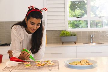 Asian woman piping green icing onto star and snowflake cookies on metal cooling rack in kitchen