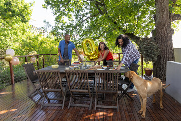 Obraz premium Diverse friends arranging party setup on wooden deck with dining table gold '0' balloon, dog